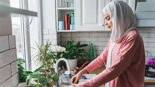 A woman wearing a pink long-sleeve shirt and a white hijab washes dishes in a modern white kitchen with a sink and small plants near the window.