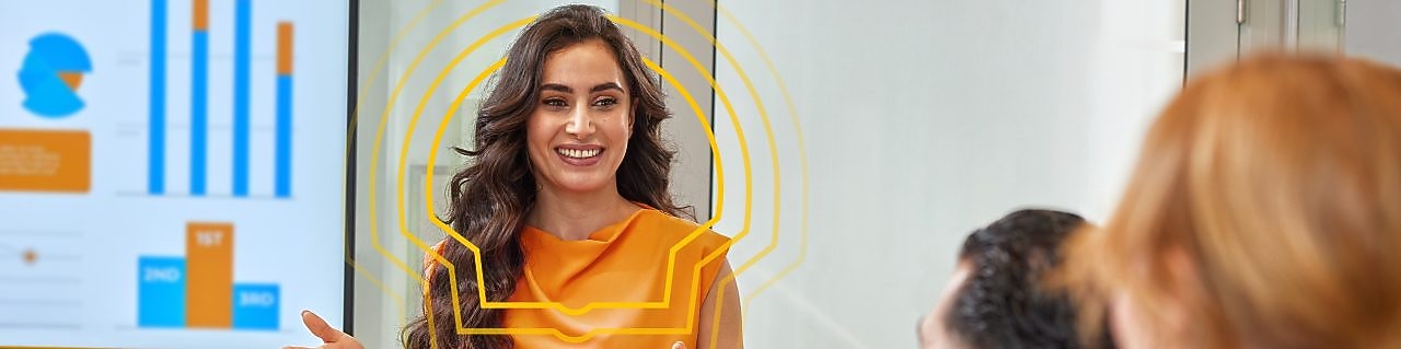 A woman with long, dark brown, curly hair presenting a report to two colleagues - one male and one female. She is wearing an orange sleeveless blouse and white pants. Behind her is a presentation deck with various graphs.