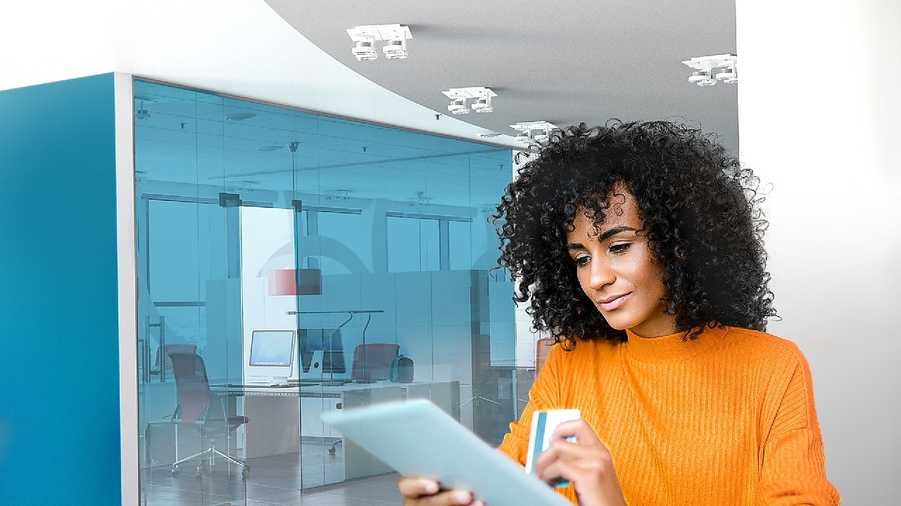 A woman with short, dark, curly hair in an orange sweater holds a tablet and a card in a modern office setting.