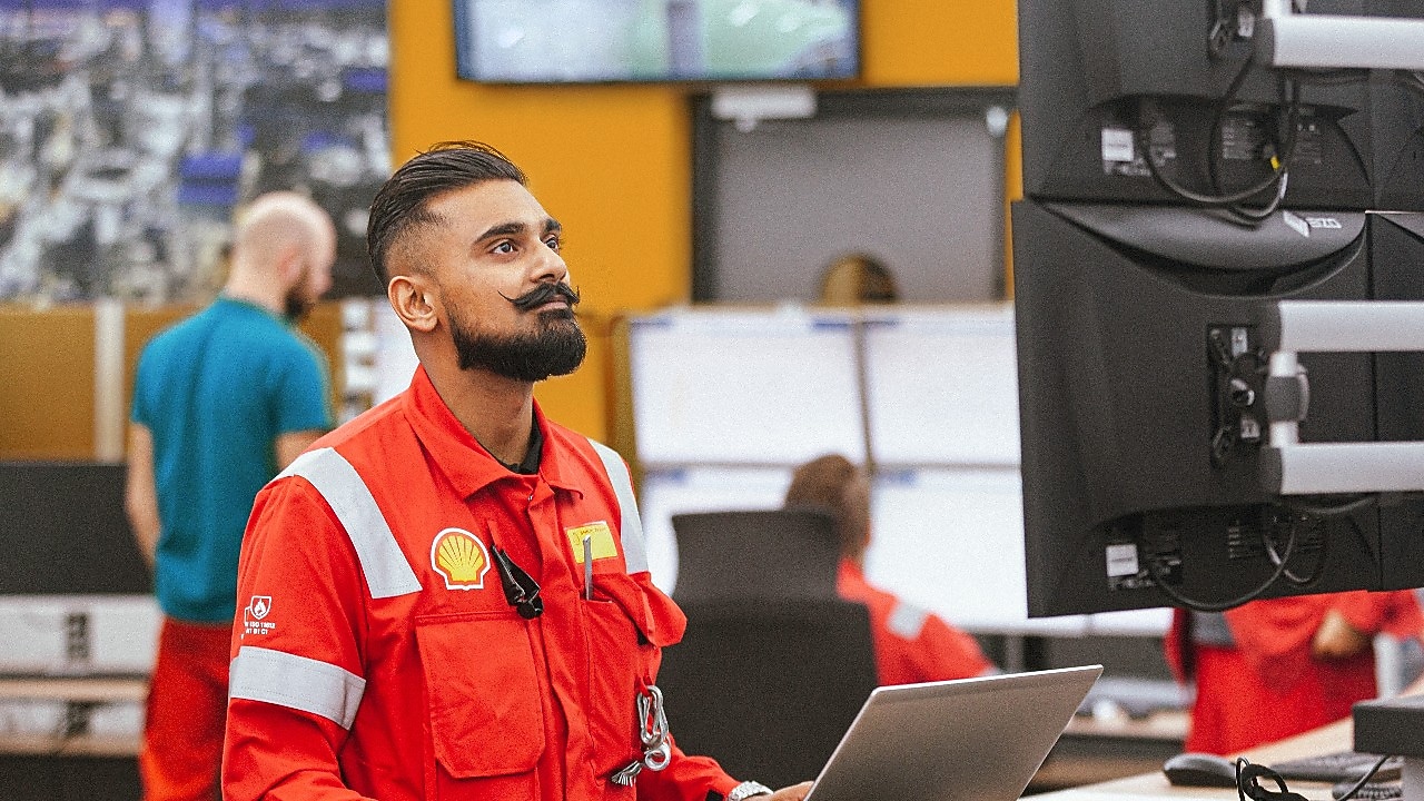 A man with dark hair wearing a Shell uniform works in front of a laptop while standing. He appears to be focused as he gazes thoughtfully at the monitor. Several computers and colleagues, also in red uniforms, are in the background.