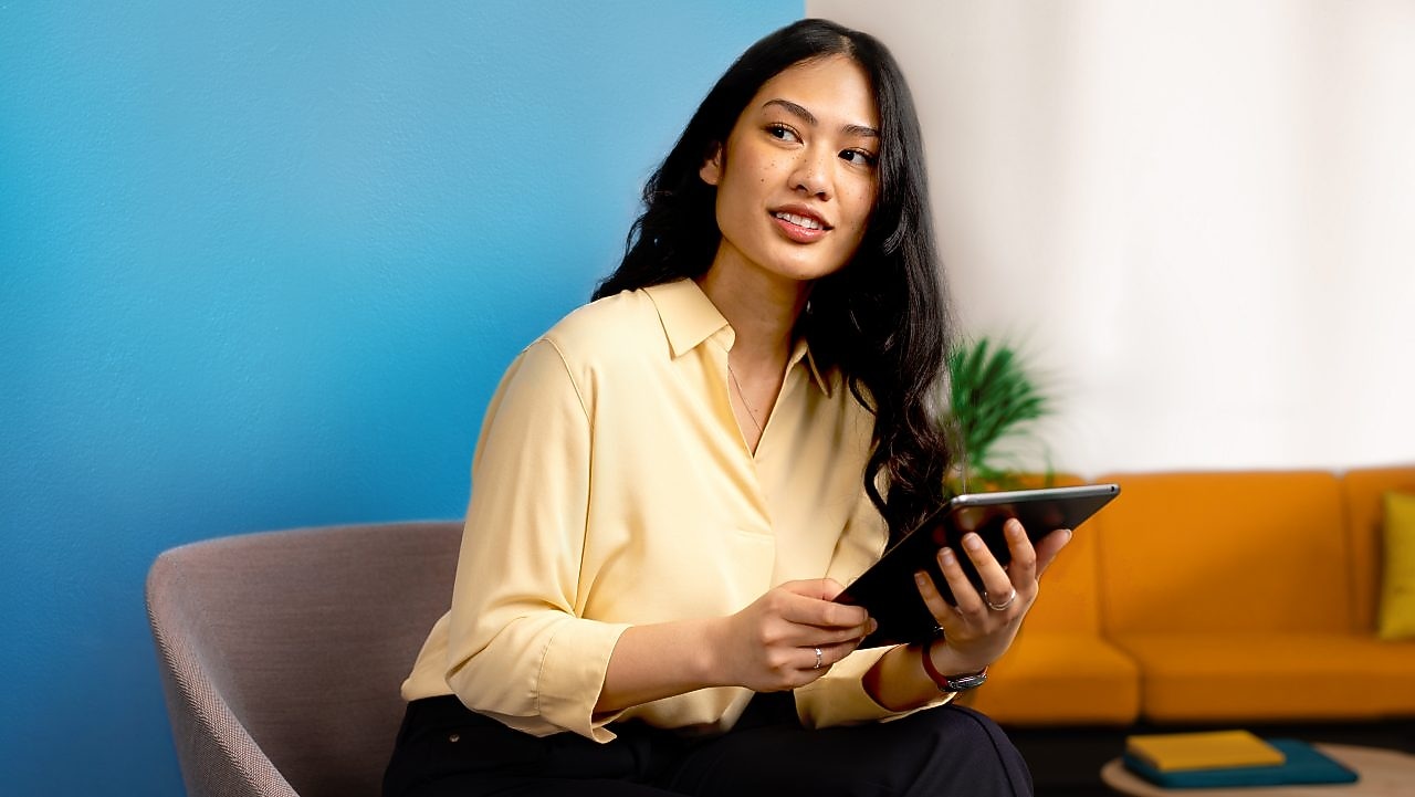 A young woman with long dark hair wearing a yellow blouse sits on a chair holding a tablet, with a blue wall and an orange sofa in the background. She is looking towards the left, smiling.