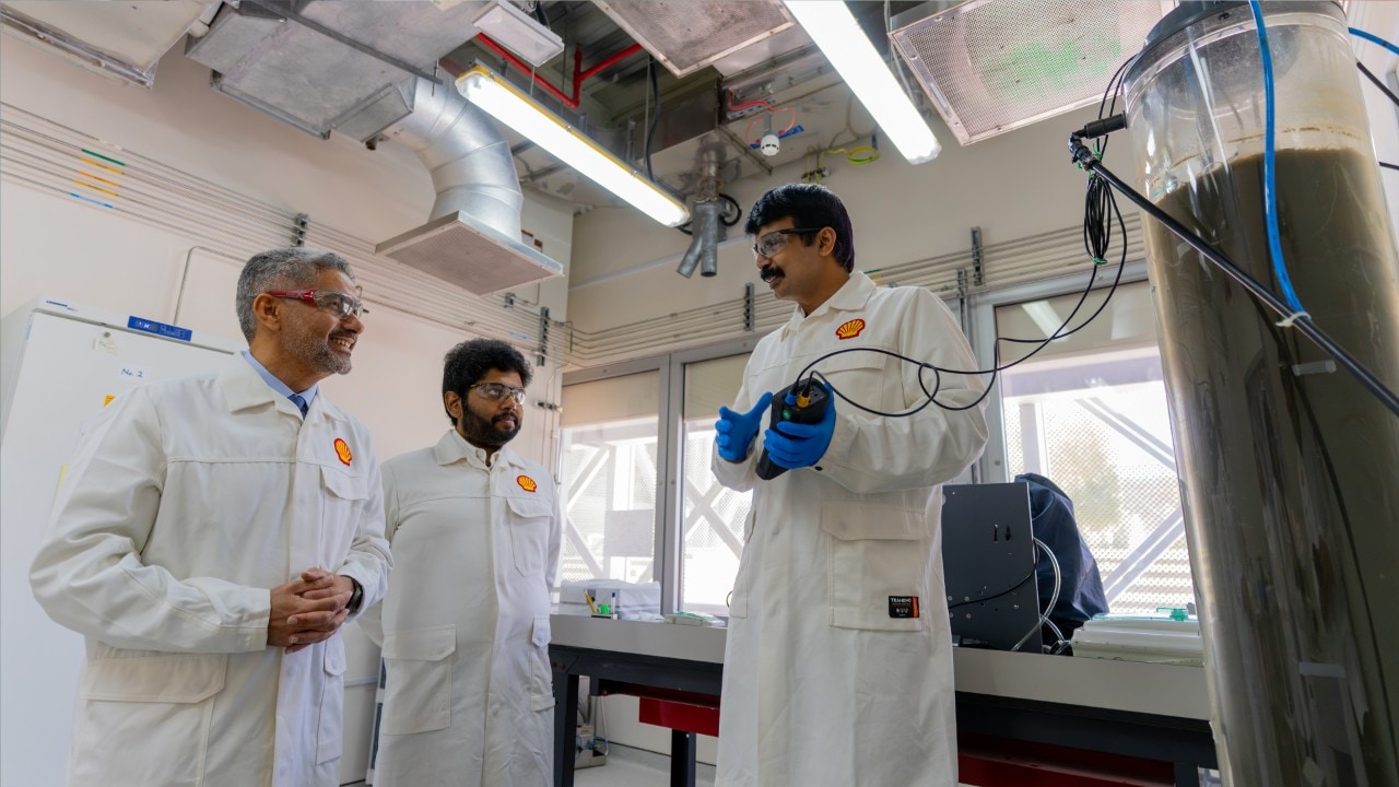 Three males wearing Shell branded laboratory coats are in conversation with one of the males holding a piece of research equipment in his hands.