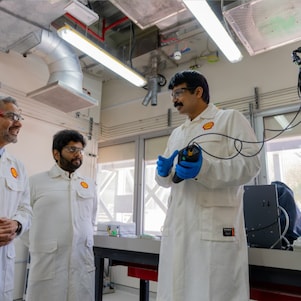 Three males wearing Shell branded laboratory coats are in conversation with one of the males holding a piece of research equipment in his hands.