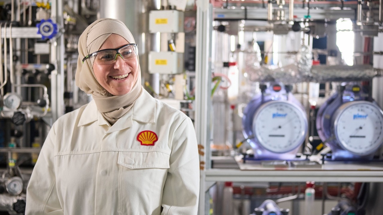 A smiling female Shell employee wearing a Shell branded laboratory coat stands in front of various laboratory equipment.