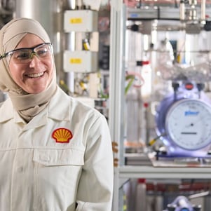 A smiling female Shell employee wearing a Shell branded laboratory coat stands in front of various laboratory equipment.