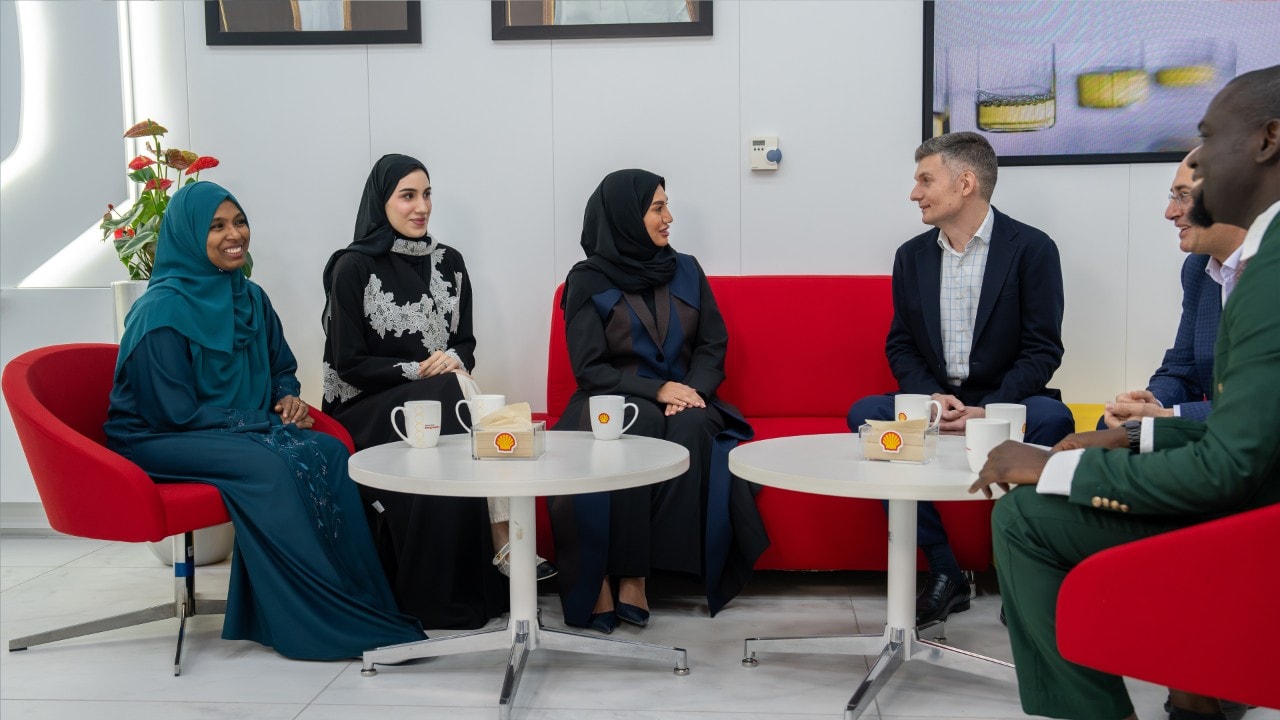 Two females and one male are sat around two coffee tables having a conversation. Shell branded mugs and cups sit on the tables.