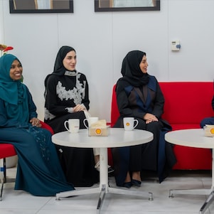 Two females and one male are sat around two coffee tables having a conversation. Shell branded mugs and cups sit on the tables.