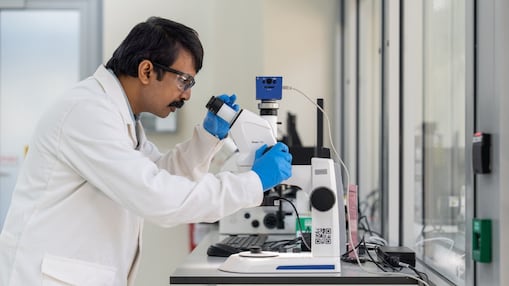 A scientist wearing a lab coat and safety glasses examines a sample using a laboratory microscope in a modern research facility.