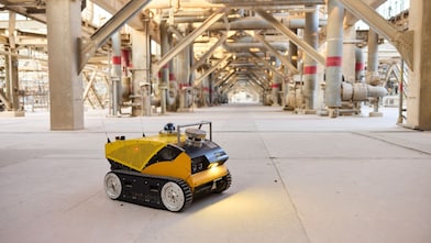 A small yellow robotic vehicle sits in the foreground of a large open space in the research facility. In the background is heavy industrial pipework.