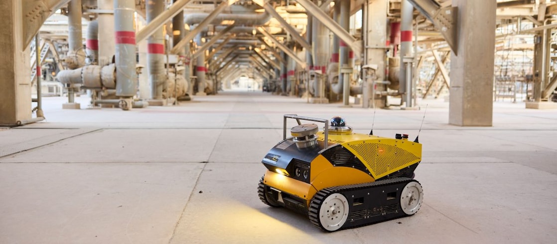 A small yellow robotic vehicle sits in the foreground of a large area full of industrial piping.