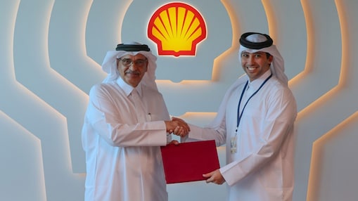 Two males are standing in front of a Shell branded wall. They are holding a red folder and shaking hands.