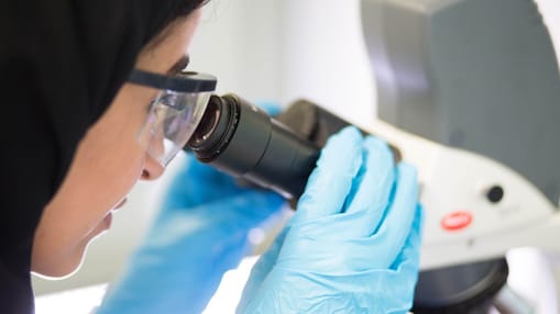 A lady wearing protective goggles and gloves using a microscope